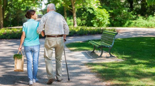 A person supporting a senior while walking outdoors in the park